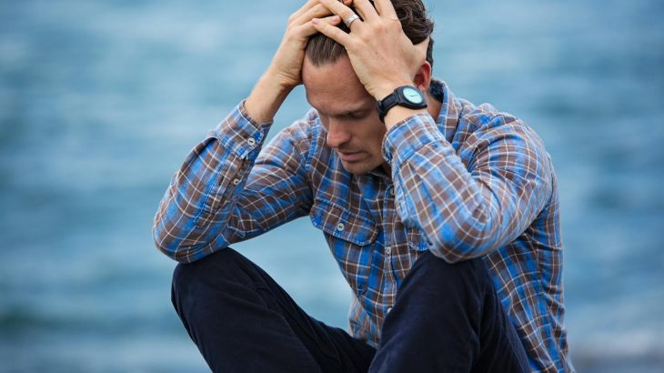 A man in a plaid shirt sits by the water looking distressed, symbolizing stress.